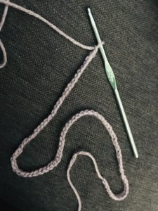 A crochet hook attached to gray yarn in the form of a simple crochet chain, all lying on a sofa cushion.