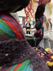 A fiber art sculpture is in the foreground, but through the sculpture there is a view of someone touching another fiber art sculpture and Anani standing before a wall display of sketchbook pages. Anani is reading from her script as she presents to the group. 